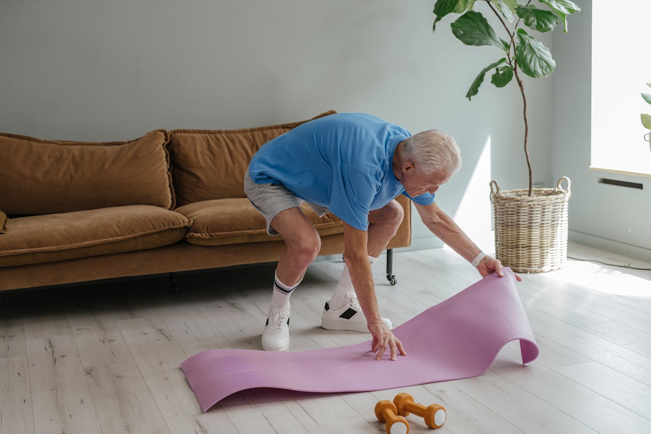 gallery-03 An elderly man sets up his yoga mat indoors, embodying a healthy lifestyle.
